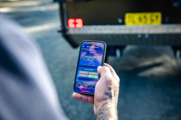 A hand holding a smartphone with the ARK Smart Connector app controlling trailer lights; the trailer’s rear and number plate are visible in the background.