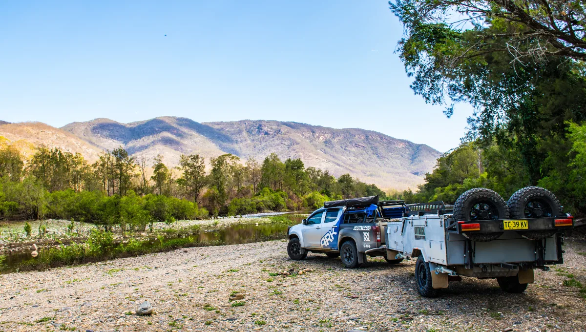 ARK-equipped 4WD ute towing an off-road camper trailer at a riverside bush camp in the mountains.